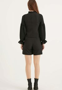 Zwarte gestructureerde top met een ruches halslijn, gecombineerd met hoge zwarte shorts. Model draagt zwarte enkellaarsjes. Gladde stof met een lichte glans.