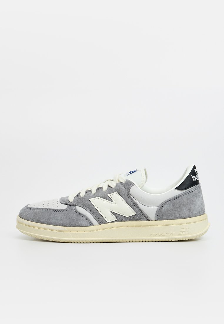 Zapatilla de ante gris y cuero blanco con puntera perforada, suela beige y acento negro en el talón que presenta el logo de "new balance".