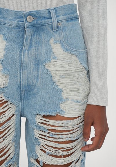 Calças de ganga de lavagem clara com rasgos pesados, apresentando bordas desfiadas e uma cintura ajustada. Fecho com botão e bolsos frontais visíveis.