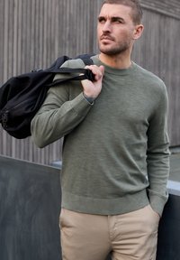 Homme en pull vert et pantalon beige portant un sac à dos noir sur l'épaule, regardant pensivement sur le côté devant un mur en bois.