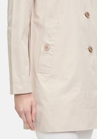 Beige leichter Mantel mit weicher Textur, ausgestattet mit zwei vorderen Taschen, doppelt geknöpft und einem geraden Saum. Taillierte Ärmel rundieren das Design.