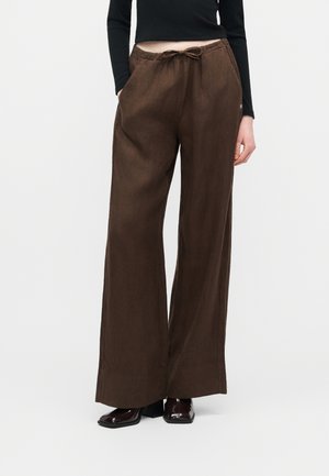 Nő, barna, magas derekú, bő szárú nadrágot visel, amelynek derékpántja húzózsinórral szabályozható, fekete hosszú ujjú felsővel és fényes sötétbarna cipővel kombinálva.