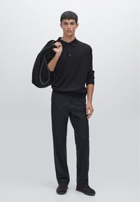 Schwarzes Strick-Polohemd mit drei Knöpfen, kombiniert mit lockeren schwarzen Hosen und dunklen Schuhen. Modell hält eine schwarze Tasche mit Trägern.