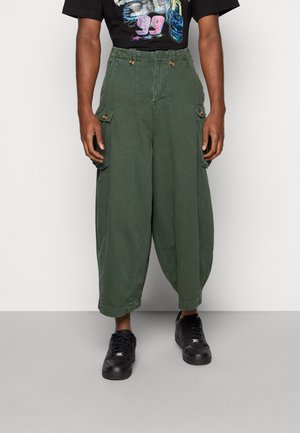 Pantaloni cargo verde oliva a vestibilità ampia con vita alta e tasche a patta con bottoni, indossati con maglietta nera con stampa grafica e scarpe da ginnastica nere.