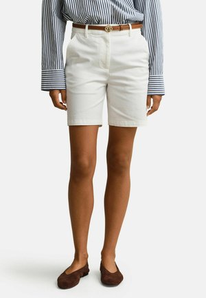 Femme portant un short blanc jusqu'aux genoux avec une ceinture marron, une chemise à manches longues rayée bleu et blanc, et des chaussures plates marron.