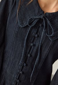Mørkt denimkjole med rundet krave, sløjfedetalje foran, lodret række af knapper og tekstureret stof med subtile sømdetaljer.