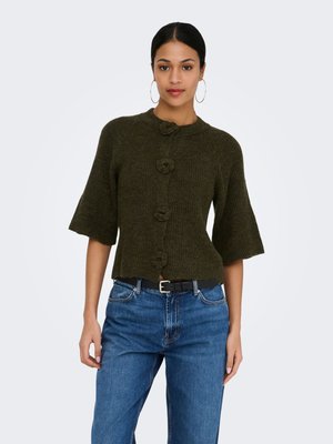 Žena oblečená v tmavozelenom krátkom pletenom kardigane s veľkými kvetinovými gombíkmi a modrých rifle s vysokým pásom a čiernym opaskom.