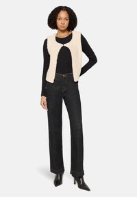 Frau steht mit einer Hand auf der Hüfte, trägt eine beigefarbene strukturierte ärmellose Weste über einem schwarzen Langarmshirt, dunkle Jeans und schwarze High Heels.