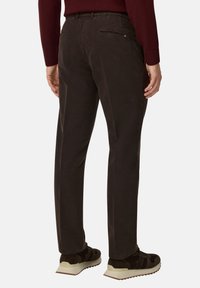 Tumši brūni bikses ar gludu tekstūru, ar aizmugurējo rāvējslēdzēja kabatu un pielāgotu piegriezumu. Kombinētas ar tumšām sporta kurpēm.