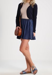 Marineblauer Cardigan, beiger gerippter Oberteil, Jeans-Skaterrock, braune Umhängetasche, graue Sandalen. Auffälliges Beintattoo auf dem linken Bein sichtbar.