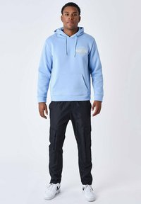 Sudadera con capucha de color azul claro, con un bolsillo frontal y un logo. Combinada con pantalones cargo negros que tienen bolsillos laterales y zapatillas blancas.