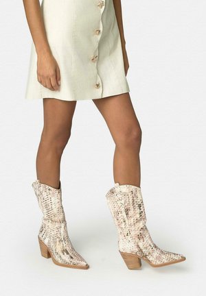 Mujer con una falda abotonada color crema y botas cowboy de media caña con estampado de serpiente en tonos claros y tacón de madera.