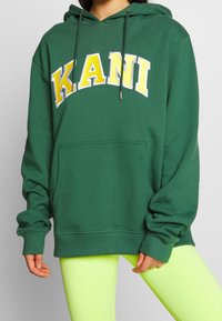 Felpa verde in tessuto con tasca a marsupio, con la scritta "KANI" in lettere bianche e gialle sulla parte anteriore. Cappuccio con laccetti.