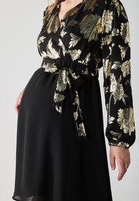 Fekete ruha arany virágmintával; sarok stílusú, hosszú ujjú és derékmegkötős. Strukturált anyag, amely szellős sziluettet biztosít.