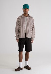 Stripet button-up skjorte i brun og hvid, sorte shorts, teal kasket, hvide sokker og brune klipklapper. Afslappet outfit med markante mønstre.