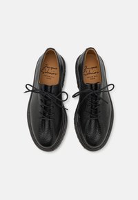 Chaussures en cuir noir avec une finition texturée, bout rond et lacets centraux. Nom de la marque visible sur la semelle intérieure beige. Semelle en caoutchouc.