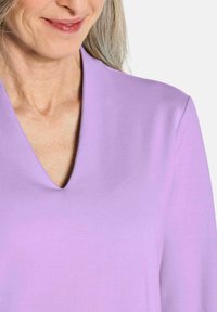 Lavendelfarbenes V-Ausschnitt-Oberteil mit glatter Textur und schlichtem Design. Der Stoff wirkt flexibel und eignet sich für die Freizeit. Keine Muster oder Verzierungen.