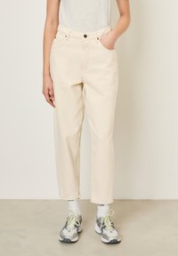 Pantalons en denim couleur crème avec une coupe droite, cinq poches et une fermeture à boutons. Associés à des chaussures de sport grises et jaunes.