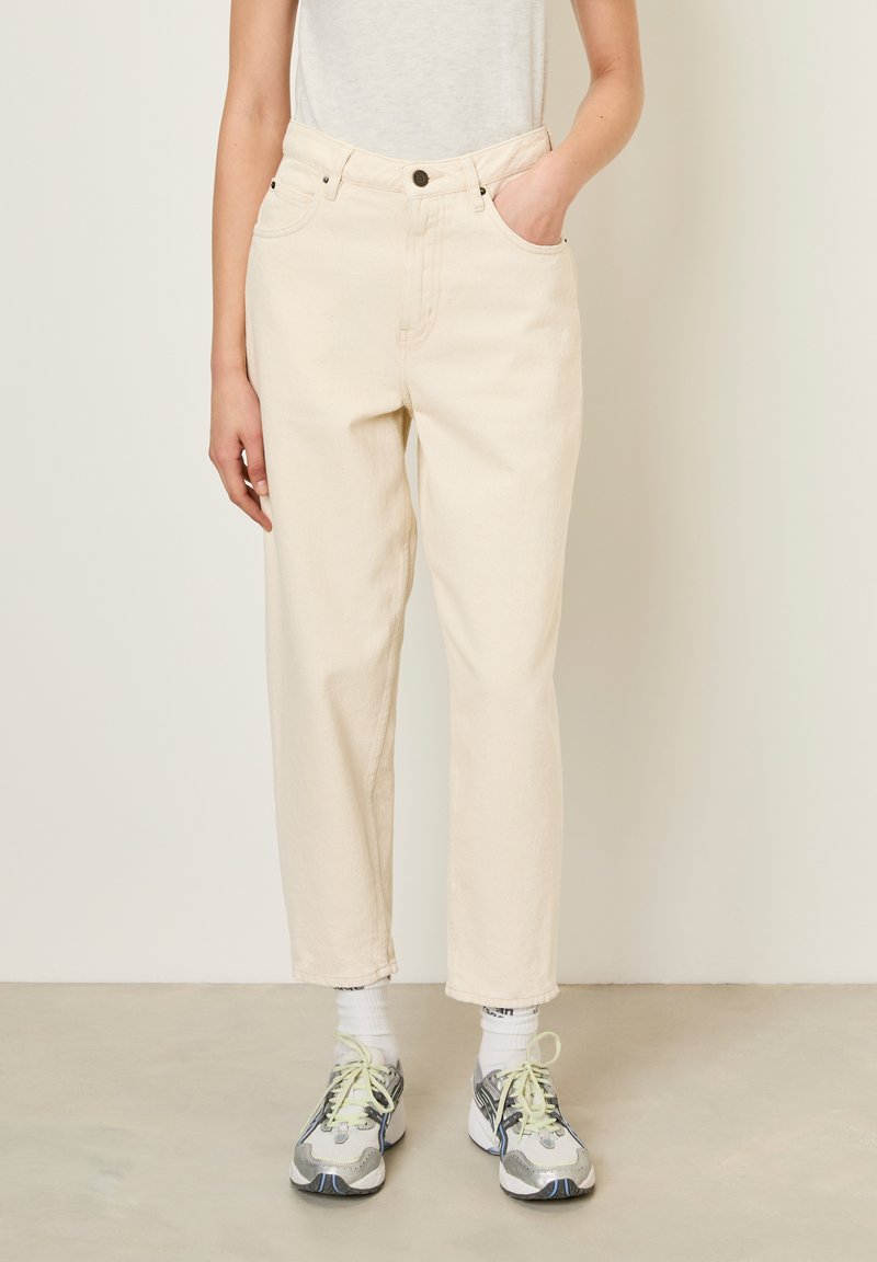Pantalons en denim couleur crème avec une coupe droite, cinq poches et une fermeture à boutons. Associés à des chaussures de sport grises et jaunes.