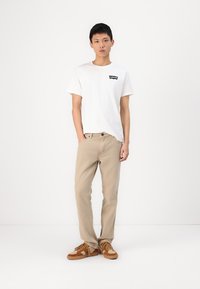 Tricou alb cu mâneci scurte și logo negru, pantaloni bej și încălțăminte maro. Ţinuta are o croială lejeră și un design simplu.