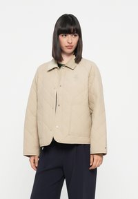 Lys beige quiltet jakke med krage, trykknapper, to sidelommer og en brodert logo på brystet. Myk stofftekstur.