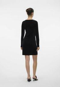 Robe en velours noir à manches longues et encolure arrondie, silhouette cintrée, longueur genou, assortie à des chaussures à talons noires.