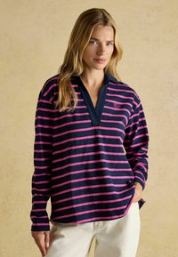 Tumepruun ja roosa triibuline pikkade varrukatega särk, millel on krae ja rinnal logo, valmistatud pehmest, tekstuurilisest kangast.