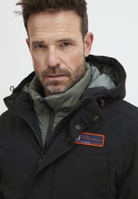 Črna zunanja jakna z zelenim notranjim podlogo, z zadrgo, kapuco in oranžno oznako na prsih. Teksturna tkanina, zasnovana za toploto.