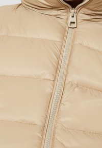 Beige vadderad jacka med hög krage, framdragkedja med kontrasterande kant och kviltad yta för isolering.