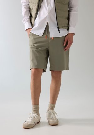 Mann trägt khakifarbene Shorts, beige Daunenweste über weißem Hemd, beige Socken und weiße Turnschuhe, steht vor einfarbigem Hintergrund.