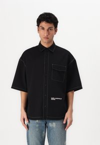 Juodas trumpomis rankovėmis marškinėliai su atleistu dizainu, kontrastinėmis siūlėmis, krūtinės kišene ir tekstu "KARL LAGERFELD JEANS" ant apatinės dalies.