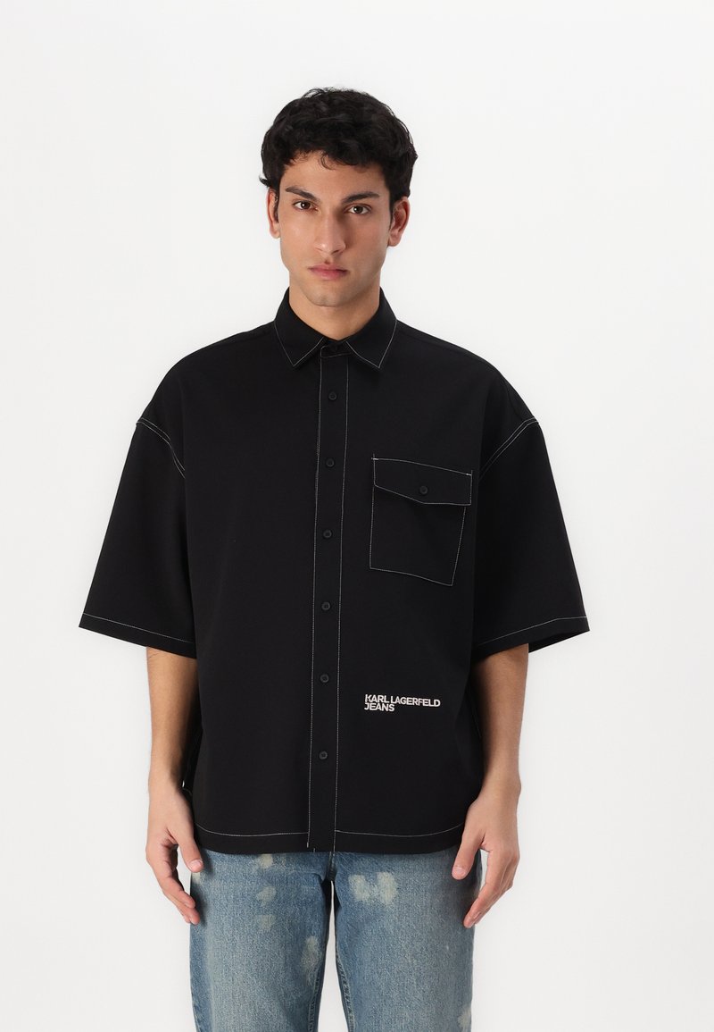 Juodas trumpomis rankovėmis marškinėliai su atleistu dizainu, kontrastinėmis siūlėmis, krūtinės kišene ir tekstu "KARL LAGERFELD JEANS" ant apatinės dalies.