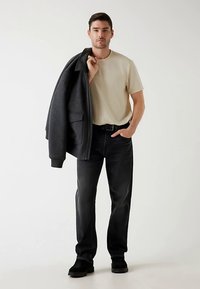 Camiseta beige, vaqueros negros y un abrigo largo de color gris oscuro. Zapatos negros, manos en los bolsillos, con una postura elegante y casual. Fondo neutro.
