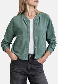 Cardigan vert texturé avec un col rond, des boutons-pression et des poignets côtelés, assorti à une chemise blanche et à un jean gris foncé.