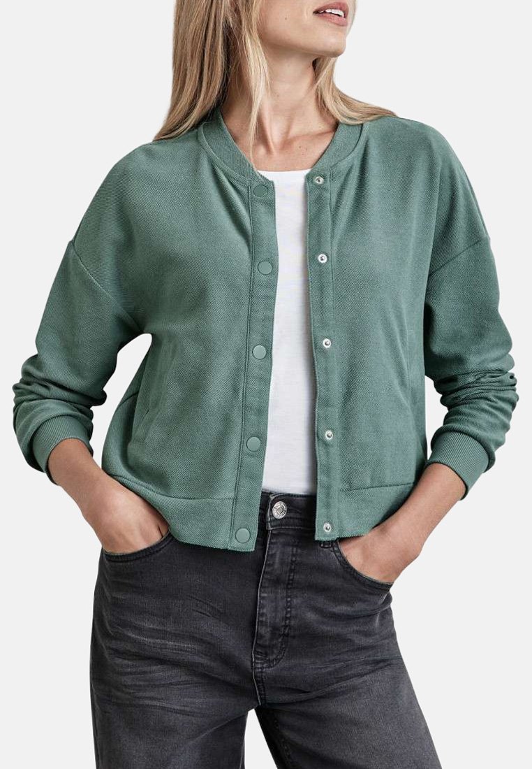 Cardigan vert texturé avec un col rond, des boutons-pression et des poignets côtelés, assorti à une chemise blanche et à un jean gris foncé.