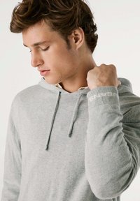Lichtgrijze hoodie van zacht materiaal, met een capuchon met trekkoord en ribgebreide boorden met een logo detail. Close-up van het kledingstuk.
