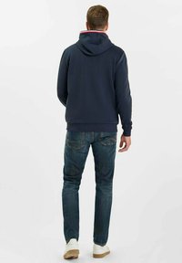 Sudadera con capucha marina con acentos en el cuello en rojo y blanco, combinada con jeans de mezclilla azul claro y zapatillas blancas, vista desde atrás.