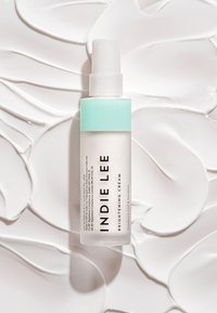 Crème éclaircissante dans un flacon translucide avec un capuchon vert menthe. L'arrière-plan présente des tourbillons de crème blanche lisse. Étiquettes de texte visibles.