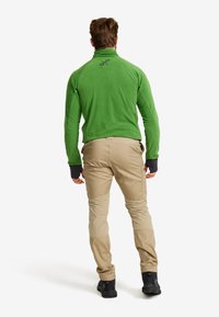 Green fleece jacket with high collar, slim fit, and black cuffs paired with beige pants featuring reinforced knees and elastic hems.