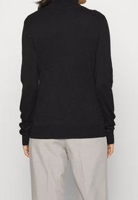 Maglione nero lavorato a maglia con collo alto, maniche lunghe e orlo a coste. Indossato sopra pantaloni grigio chiaro, caratterizzati da una texture liscia.