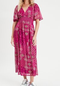Lang rosa kjole med blomster- og geometriske mønstre, korte flagrende ærmer, en dyb V-udskæring og en smocket talje. Båret med grå og hvide sneakers.