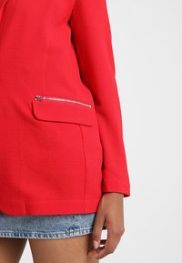 Blazer texturé rouge avec poche zippée en argent au niveau de la taille, porté avec une jupe en denim, montrant le bras droit et la main détendue sur le côté.