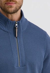 Blauer Strickpullover mit hohem Kragen, ausgestattet mit einem Reißverschluss auf der Vorderseite und einem gestreiften Zipp-Band. Strukturierter Stoff und gerippter Kragen.