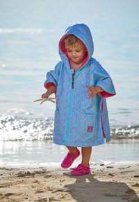 Blå poncho med mønster af pink flamingoer, der har hætte, frontlynlås og en lomme. Barnet har pink sko på og står på en sandstrand.