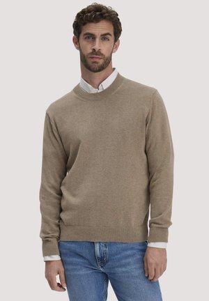 Mann mit lockigem Haar und Bart trägt einen beigen Pullover über einem weißen Hemd und blauen Jeans, steht vor hellem einfarbigem Hintergrund.