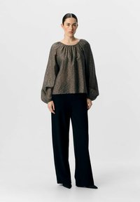 Blouse marron à motifs avec des manches bouffantes, col froncé, associée à un pantalon large noir et des chaussures pointues noires. Style minimaliste.