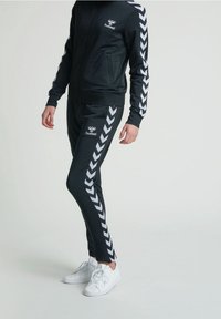 Black tracksuit with white chevron stripes on sleeves and sides, featuring a zippered jacket and tapered pants. Paired with white sneakers.