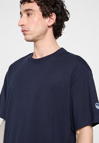 Camiseta de cuello redondo azul marino hecha de tejido texturizado. Presenta un corte relajado y un pequeño parche con el logo en la mangas izquierda.