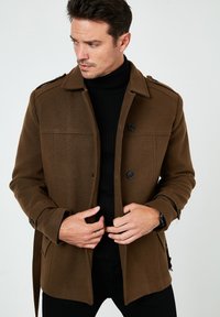 Abrigo marrón de mezcla de lana con cierre de botón, hombreras y bolsillos laterales, utilizado sobre un jersey de cuello alto negro. Textura suave, ajuste a medida.