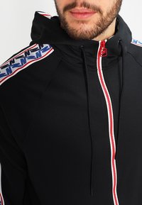 Svart zip-up huvtröja med röd dragkedja och snören, med vita och blå mönstrade detaljer på axlarna. Mjuk tygstruktur.